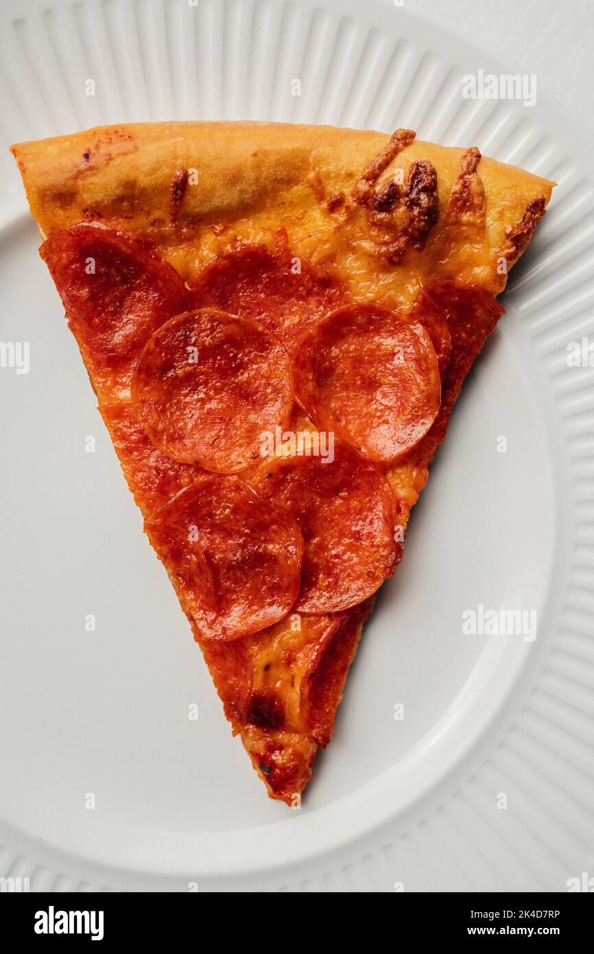 Draufsicht auf eine einzelne Scheibe Pepperoni-Pizza auf einer weißen Keramikplatte, die wie ein Pappteller aussieht Stockfoto
