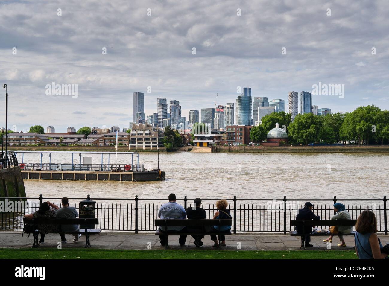 An einem warmen späten Frühlingstag sitzen einige Leute auf den Bänken entlang der Themse in Greenwich. Im Hintergrund Canary Wharf Skyline Landschaft. Stockfoto