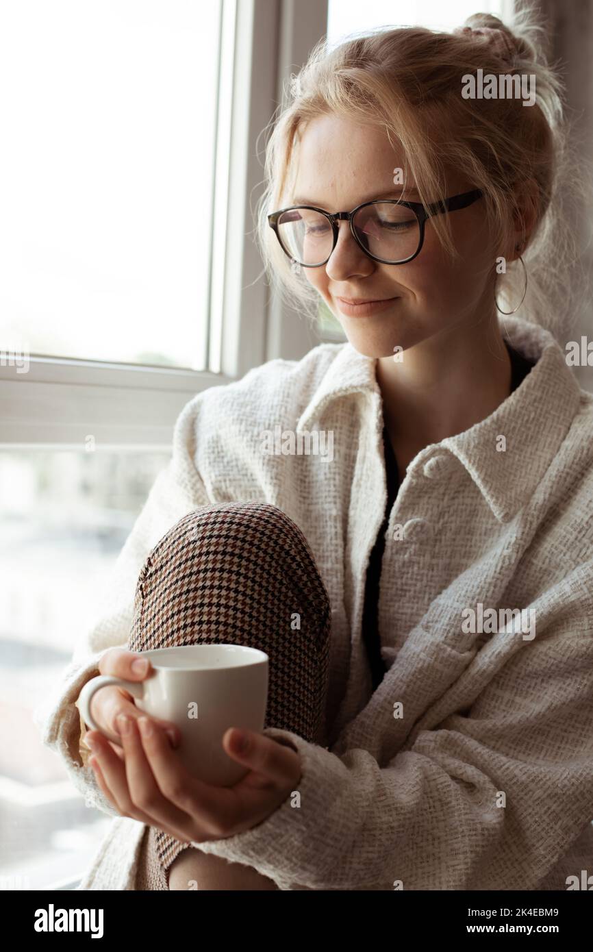 Vertikal Moderne, nachdenklich lächelnde blonde Frau in Brillen und warmer Kleidung sitzt auf dem Fenster und hält ein heißes Getränk. Zen-ähnlich Stockfoto