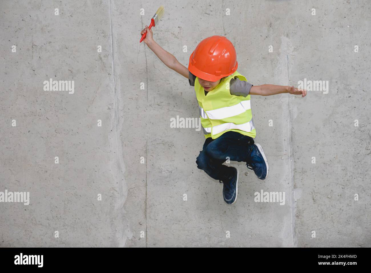Junge mit Hardhut und Sicherheitsweste, der vor einer Betonwand springt Stockfoto