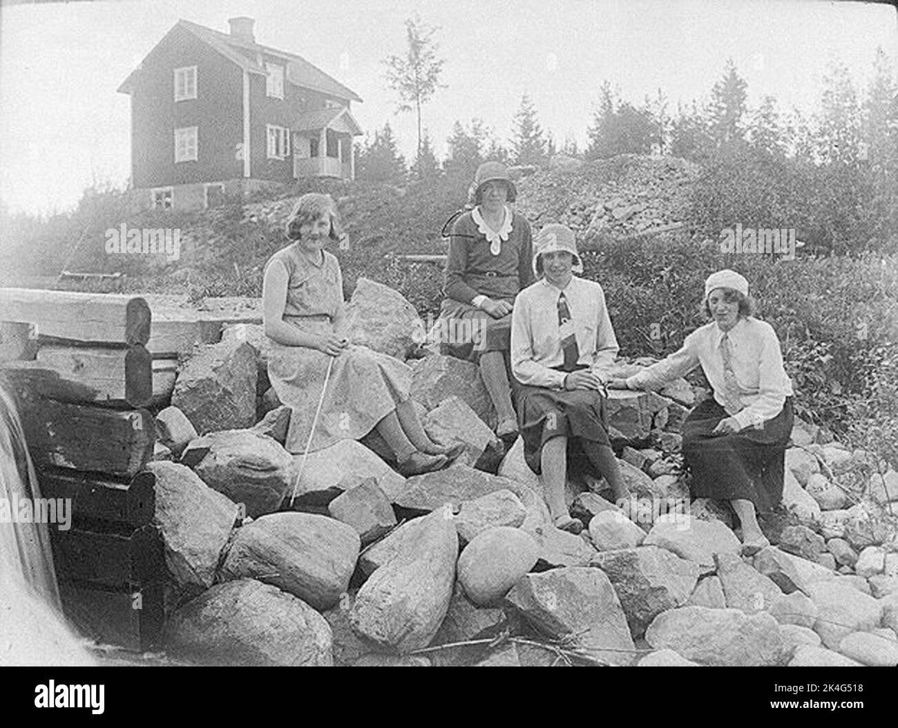 Vier Frauen sitzen auf einem Steinhaufen, ein Haus im Hintergrund. Nordisch Stockfoto