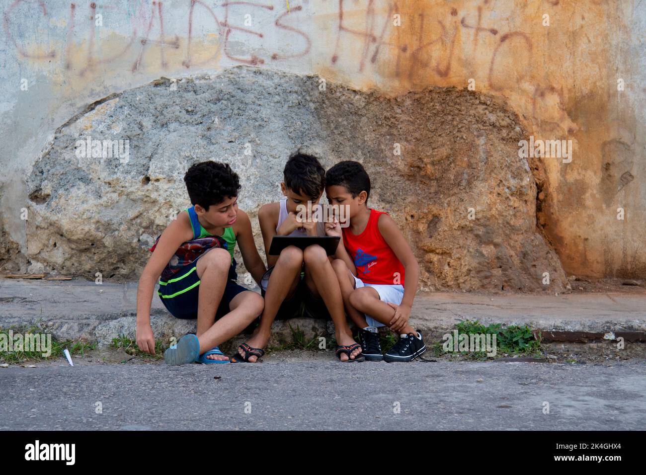 Drei junge kubanische Jungen, die in Havanna, Kuba, auf einem Bürgersteig auf einem kleinen Computer sitzen und genau hinschauen. Stockfoto