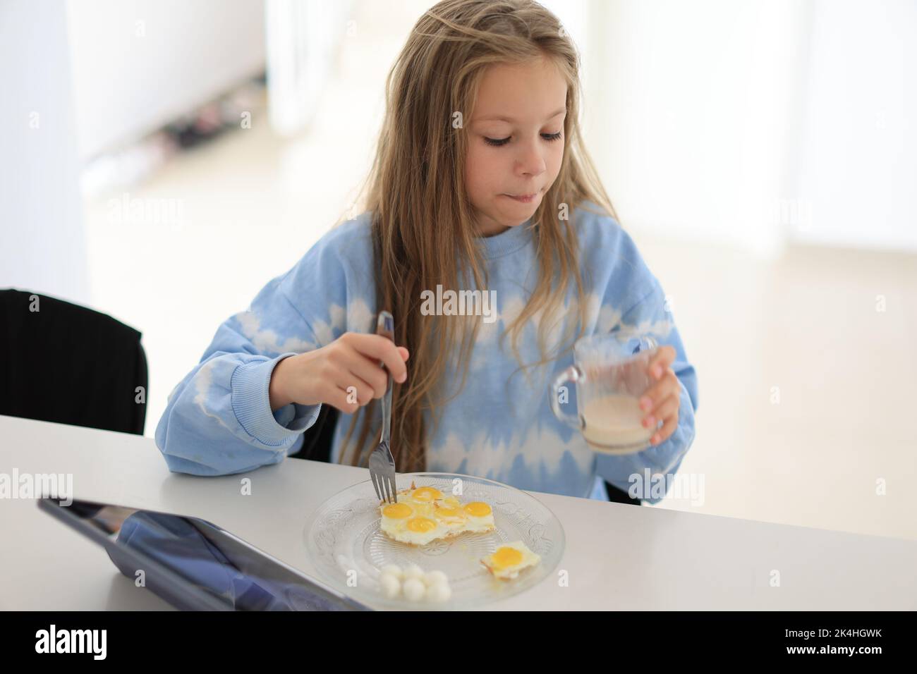 Das kleine Mädchen trinkt am Morgen die Milch der ganzen Kuh mit dem Ei und sitzt am Tisch. Richtige Ernährung für Kinder Stockfoto