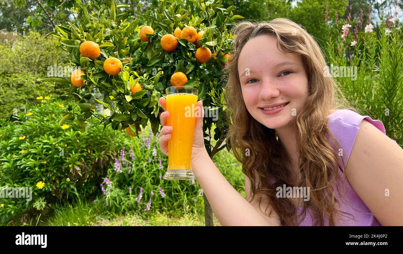 Ein Teenager-Mädchen steht mit einem Glas Orangensaft auf dem Hintergrund eines Zitrusbaums Mandarinen oder Orangen hängen an einem Baum. Sie lächelt und schaut in den Rahmen kann verwendet werden, um Säfte zu werben. Stockfoto
