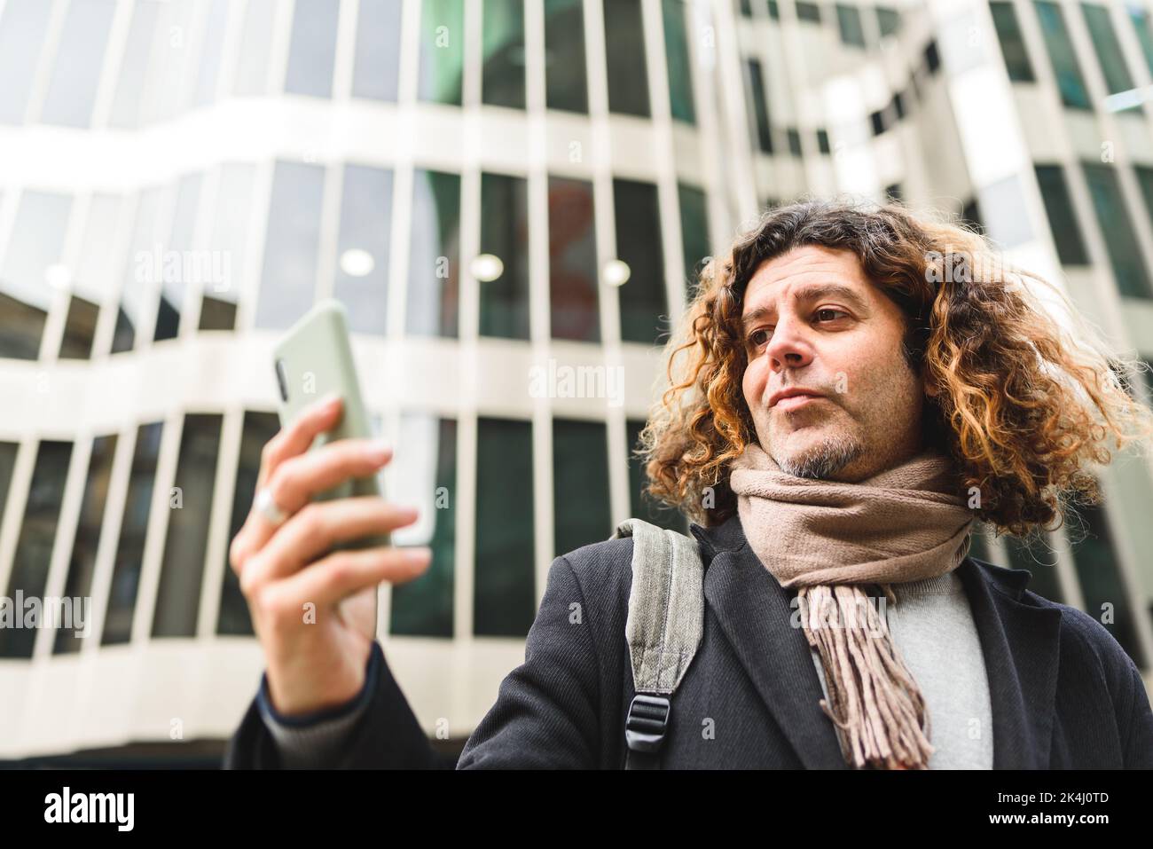 Von unten selbstsicherer reifer Mann mit lockigen Haaren, der Nachricht auf dem Handy liest, während er gegen ein modernes Gebäude in der Innenstadt steht Stockfoto