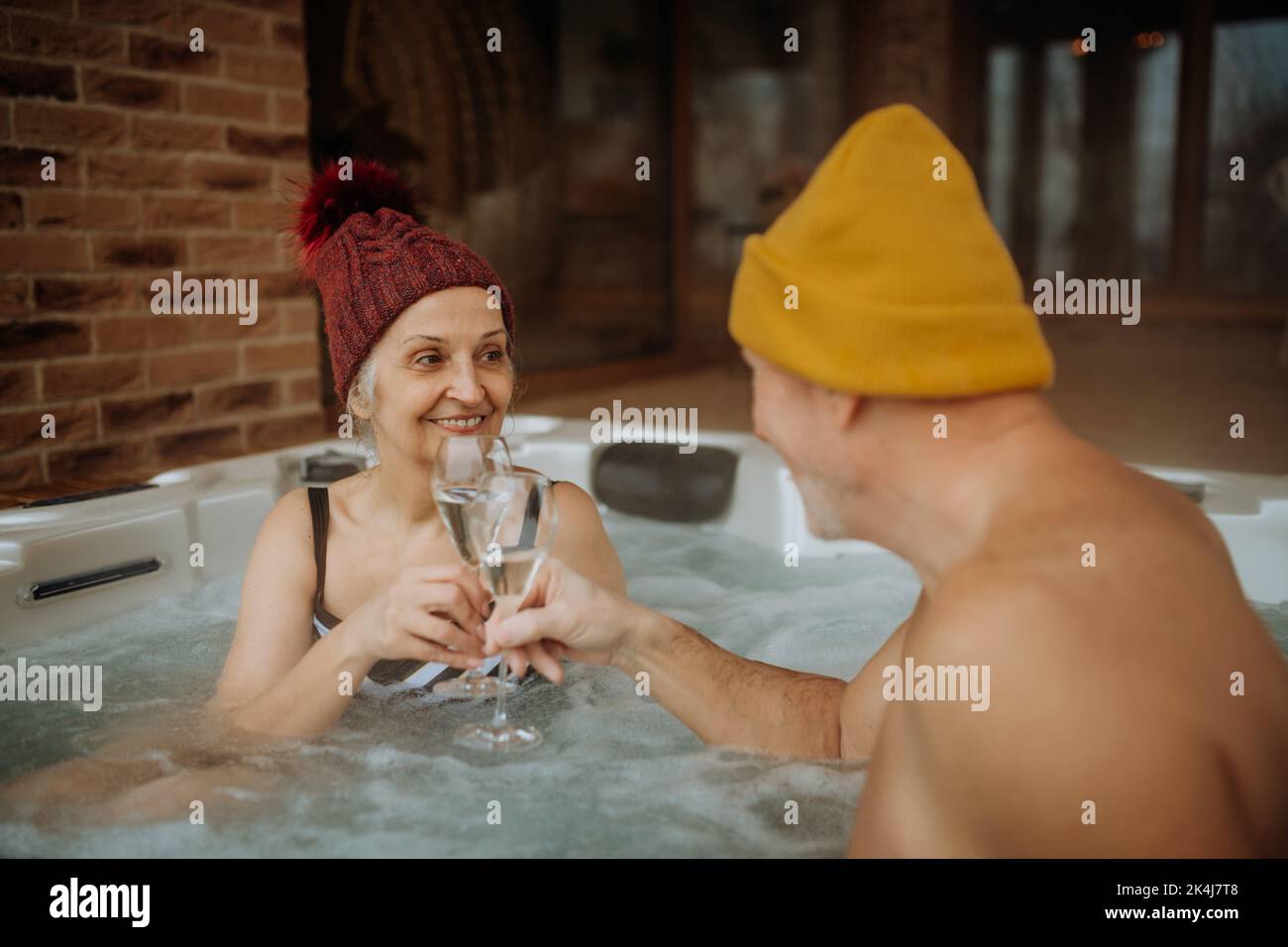 Ein Seniorenpaar mit geknickter Kappe genießt an kalten Wintertagen gemeinsam eine Badewanne im Freien und klirrende Gläser auf der Terrasse. Stockfoto