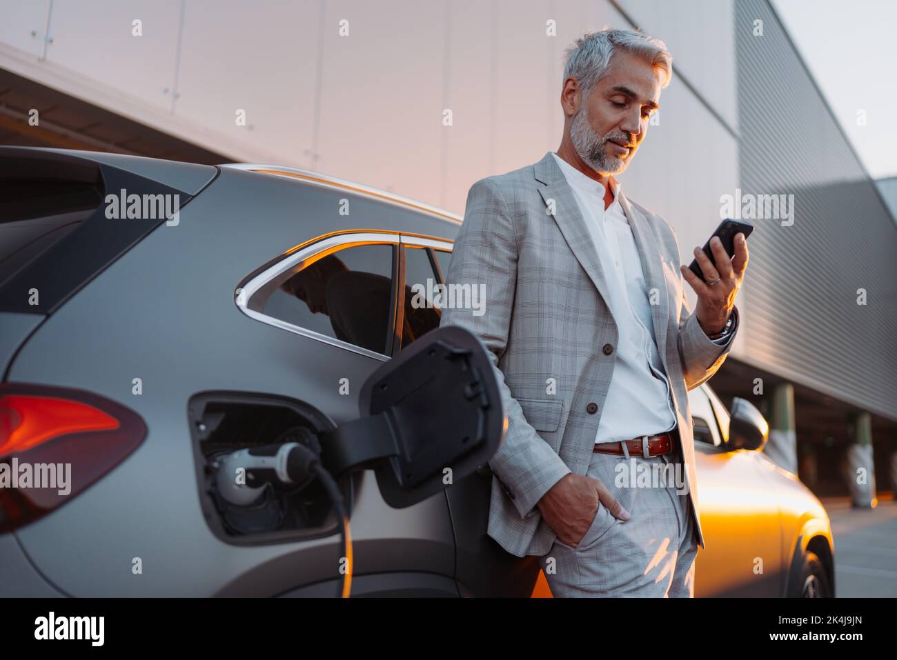 Geschäftsmann hält Smartphone, während er das Auto an der Ladestation für Elektrofahrzeuge lädt, Nahaufnahme. Stockfoto