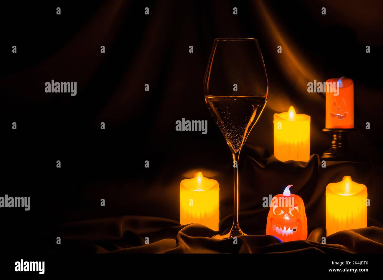 Ein Glas Weißwein mit kleiner halloween Laterne und verschwommener Fokuskerze auf dunklem Stoffhintergrund. Halloween Dinner Party Konzept. Stockfoto