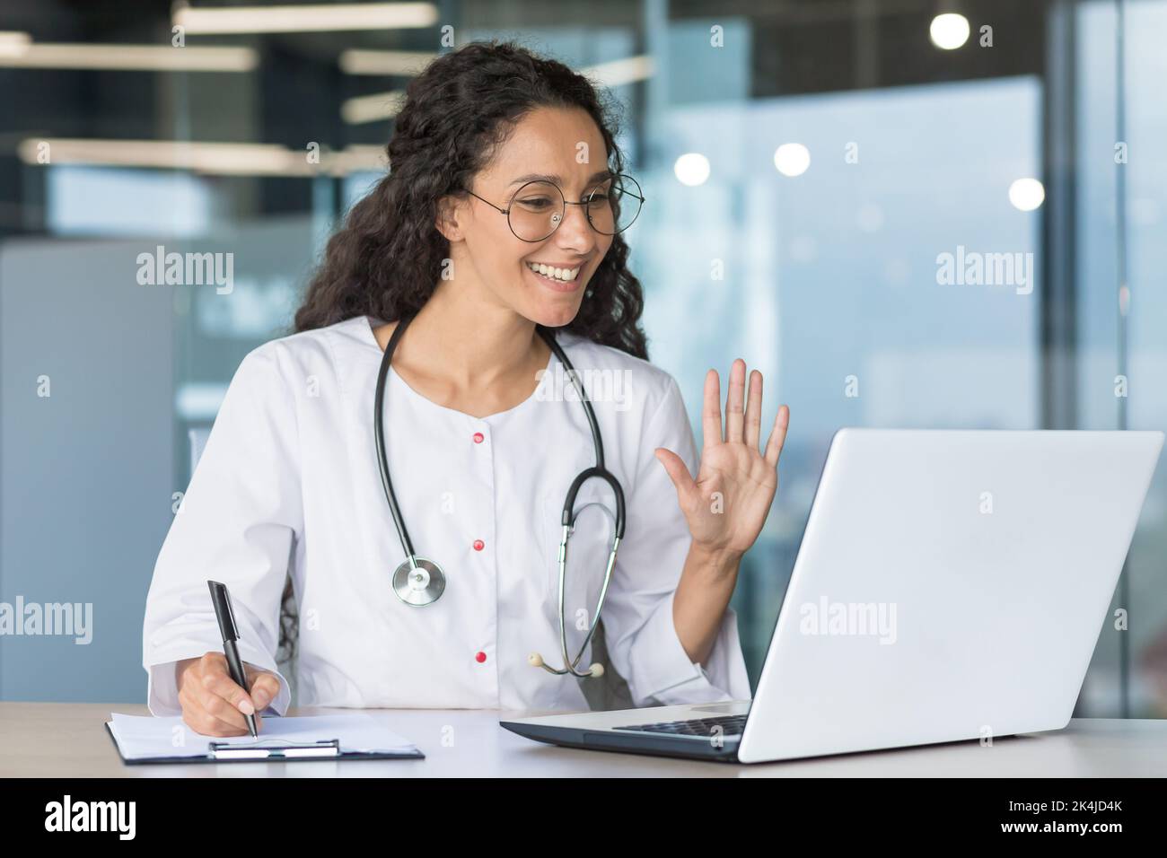 Der Arzt ist online. Eine junge Ärztin aus Lateinamerika nimmt an einer Online-medizinischen Konferenz Teil. Er winkt mit der Hand auf die Laptop-Kamera, sagt Hallo, lächelt, macht Notizen. Stockfoto