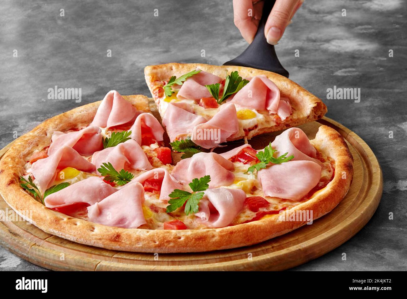 Weibliche Hand mit Servierspatel, Pizza mit geschmolzenem Käse, Schinken, Wachteleiern, Tomaten und Gemüse Stockfoto