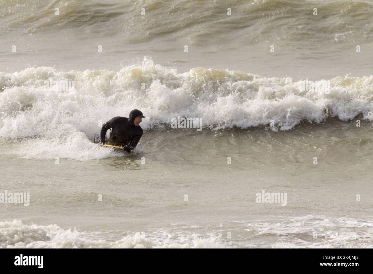 Bodysurfer im Meer auf dem Surfbrett in nassem (oder trockenem) Anzug, der unter windigen Bedingungen mit großen Wellen und weißen Wellen, die an den Strand krachen, die Brandung reitet. Stockfoto