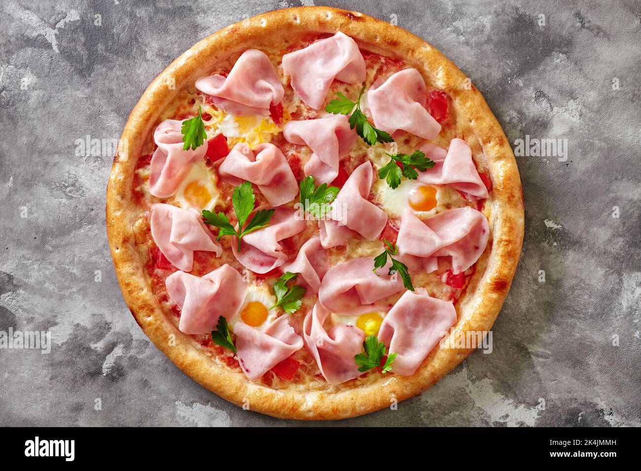 Blick von oben auf Pizza mit Schinken, Wachteleiern, Tomaten, Mozzarella und Grüns auf grauem Steingrund Stockfoto
