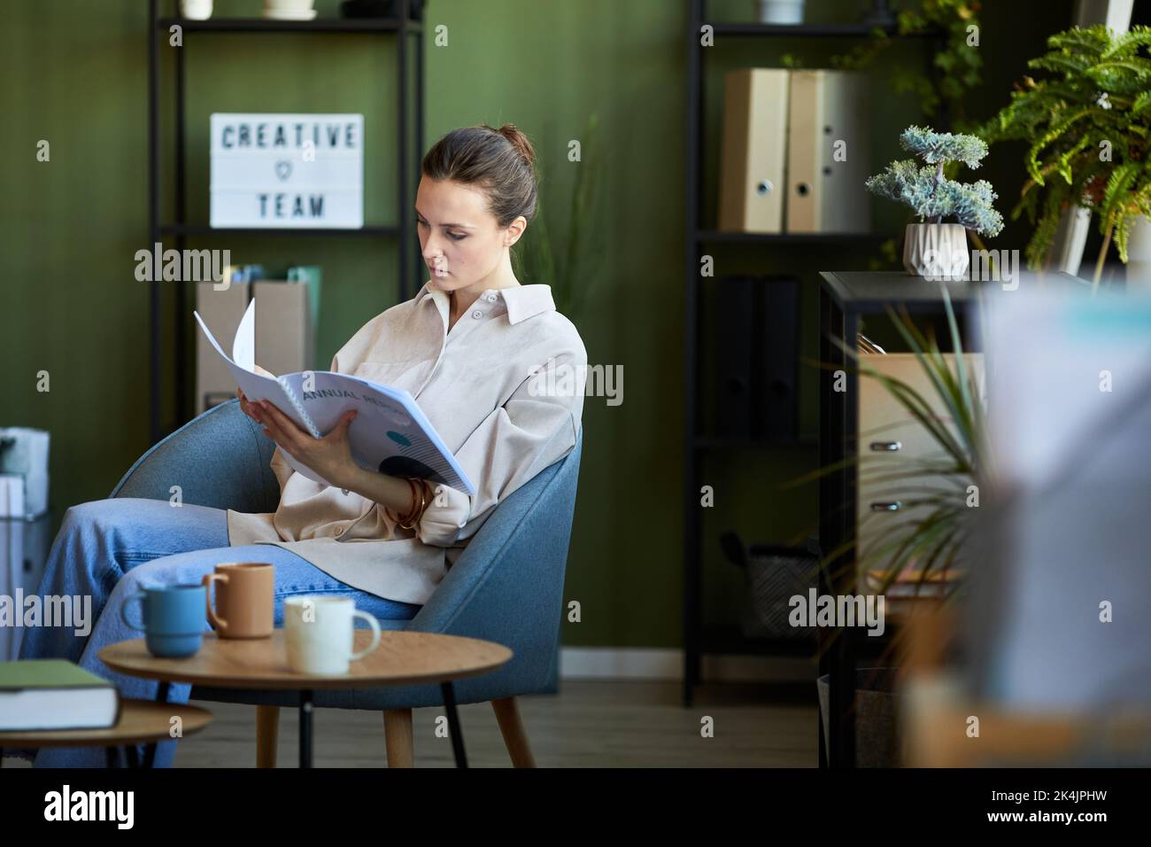Junge Manager oder Ökonom in Casualwear sitzen in einem bequemen Sessel und schauen durch Handbuch im Büro im Freiraum Stockfoto