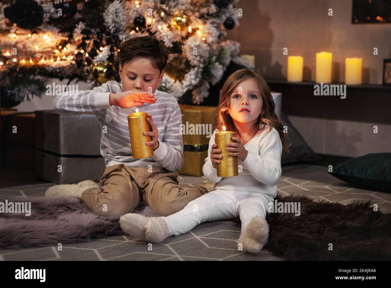 Entzückende Kinder, Junge und Mädchen halten gelbe Kerzen und machen Wunsch. Bruder und nette Schwester sitzen auf dem Boden in dunklen Raum. Weihnachtsbaum und Präs Stockfoto