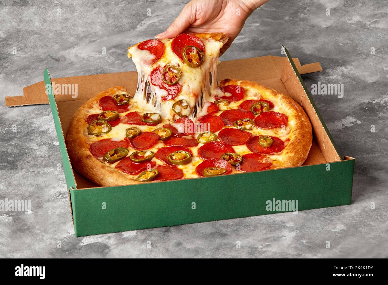 Weibliche Hand nehmen Scheibe Pizza mit geschmolzenem Mozzarella, Pepperoni und Jalapeno aus Karton Stockfoto
