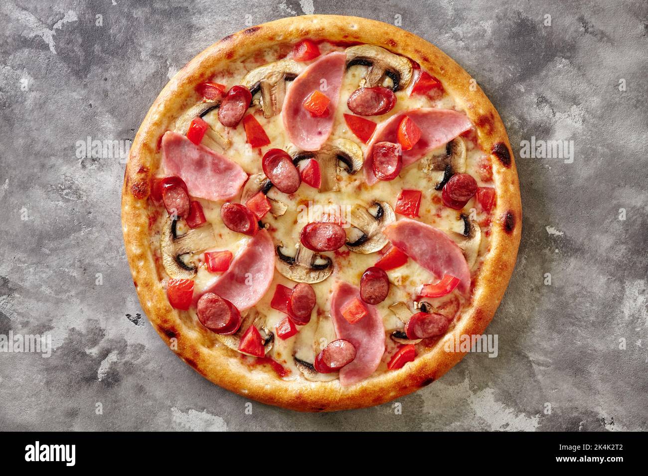 Frisch gebackene Pizza mit Jagdwürsten, Schinken, Pilzen und Tomaten auf grauer Steinoberfläche Stockfoto