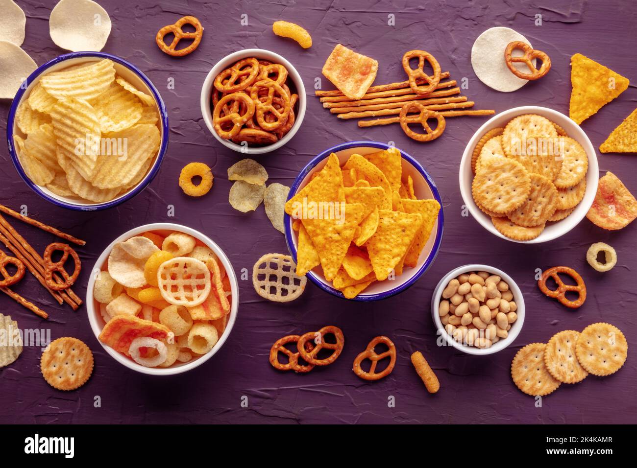 Salzige Snacks. Party-Food-Mix. Kartoffel- und Tortilla-Chips, Cracker und andere Vorspeisen in Schüsseln, über dem Kopf flach auf einem violetten Hintergrund geschossen Stockfoto