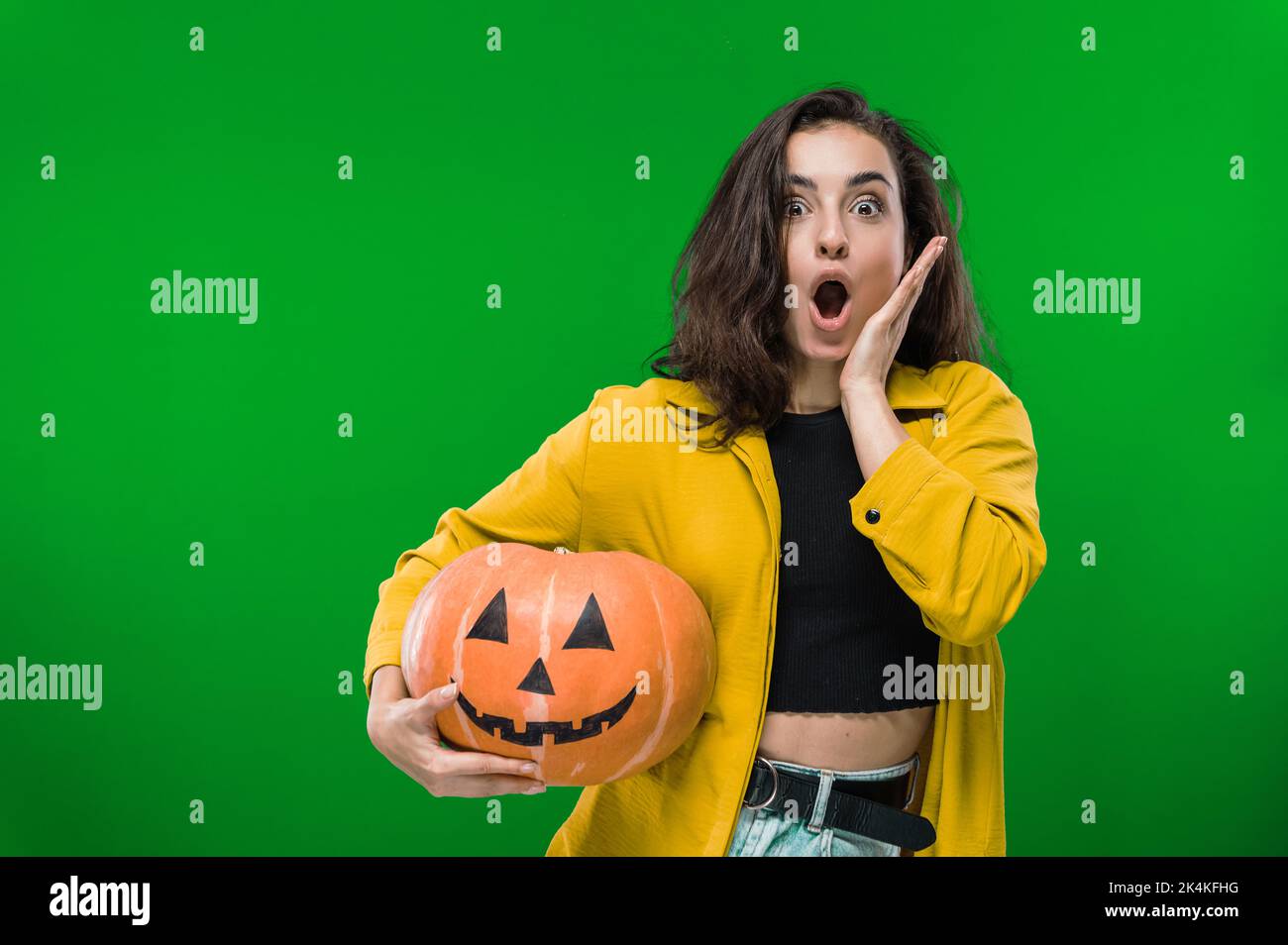 Erschrocken aufgeregt schockierte kaukasische Frau in legerer Kleidung, hält gemalten halloween-Kürbis, steht auf isoliertem grünen Hintergrund, Blick auf die Kamera in Staunen. Halloween-Party, Albtraum-Konzept Stockfoto
