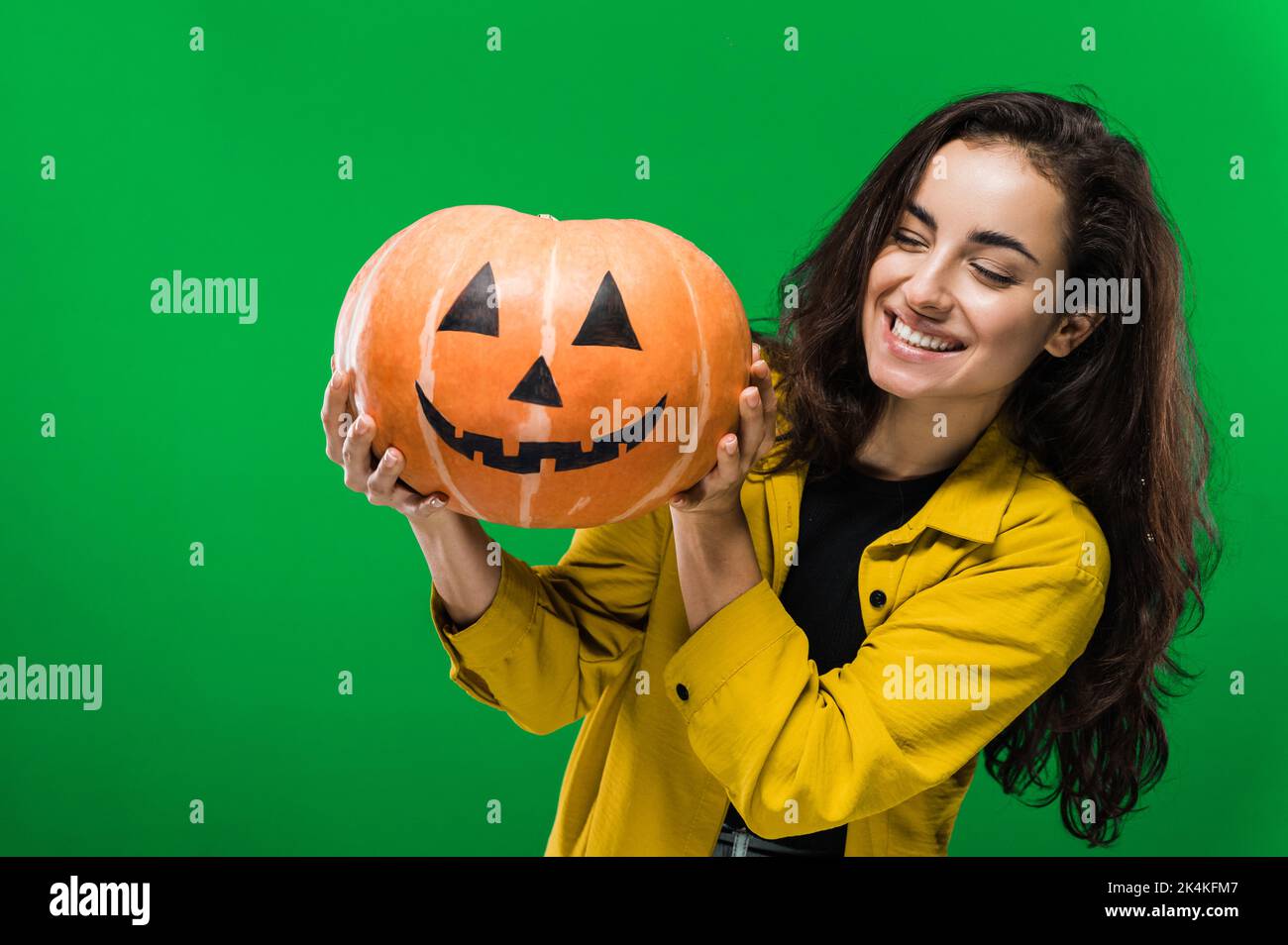 Foto von einer fröhlichen kaukasischen Frau in legerer Kleidung, mit gemaltem halloween-Kürbis, stehend auf isoliertem grünem Hintergrund, lächelnd. Halloween-Party, Albtraum-Konzept Stockfoto
