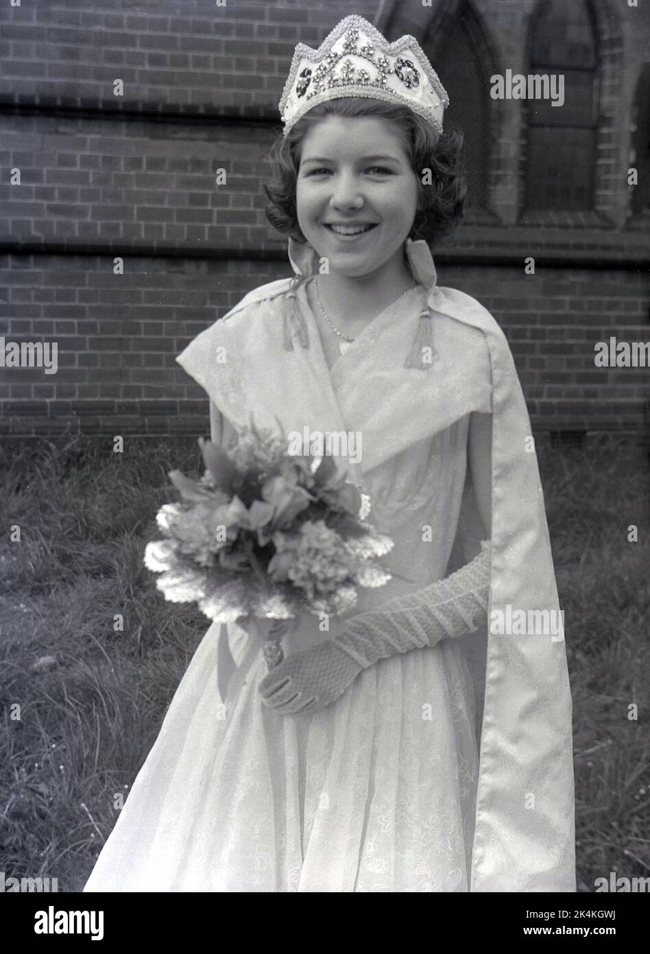 1959, historisch, ein Teenager-Mädchen, die lokale May Queen, steht für ihr Foto in ihrem langen Kleid, mit Blumen und Krone auf dem Kopf, Leeds, England, Großbritannien. Ihr Foto wird auf dem Gelände der Kirche vor dem Karneval gemacht, wo sie eine prominente Figur bei den traditionellen Maifeierlichkeiten sein wird, einem alten europäischen Festival, das den Beginn des Sommers markiert. Stockfoto