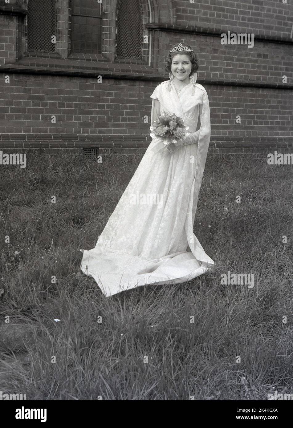 1959, historisch, ein Teenager-Mädchen, die lokale May Queen, steht für ihr Foto in ihrem langen Kleid, mit Blumen und Krone auf dem Kopf, Leeds, England, Großbritannien. Ihr Foto wird auf dem Gelände der Kirche vor dem Karneval gemacht, wo sie eine prominente Figur bei den traditionellen Maifeierlichkeiten sein wird, einem alten europäischen Festival, das den Beginn des Sommers markiert. Stockfoto