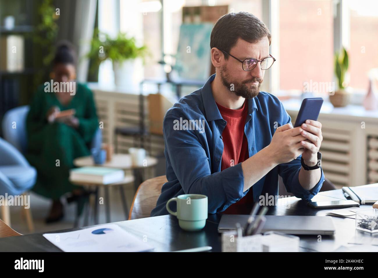 Junger seriöser Geschäftsmann in Brillen und Casualwear Scrollen im Smartphone, während er nach Online-Daten sucht oder Videos ansieht Stockfoto