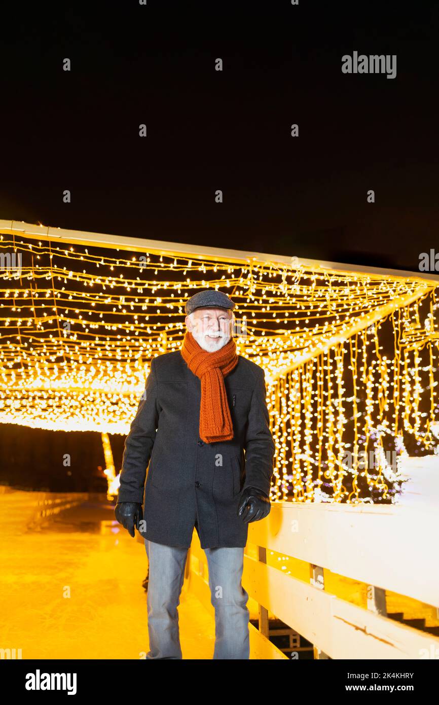 Glücklicher älterer Mann mit Schal und Jacke unter den Weihnachtslichtern lächelnd Stockfoto