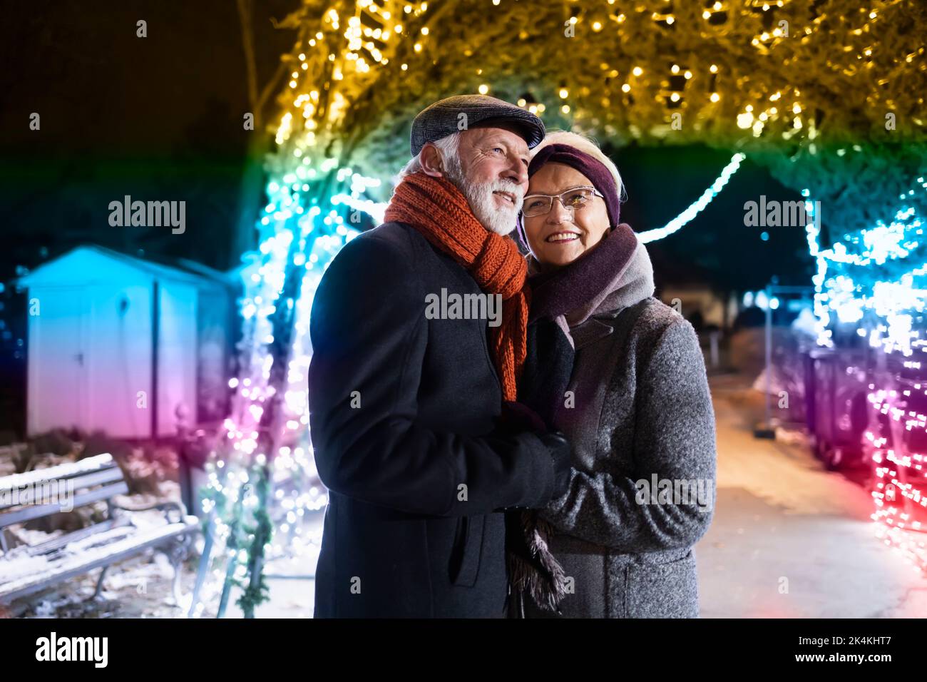 Ältere Paare lächeln in liebevoller Umarmung im Winterwunderland mit Weihnachtslichtern Stockfoto