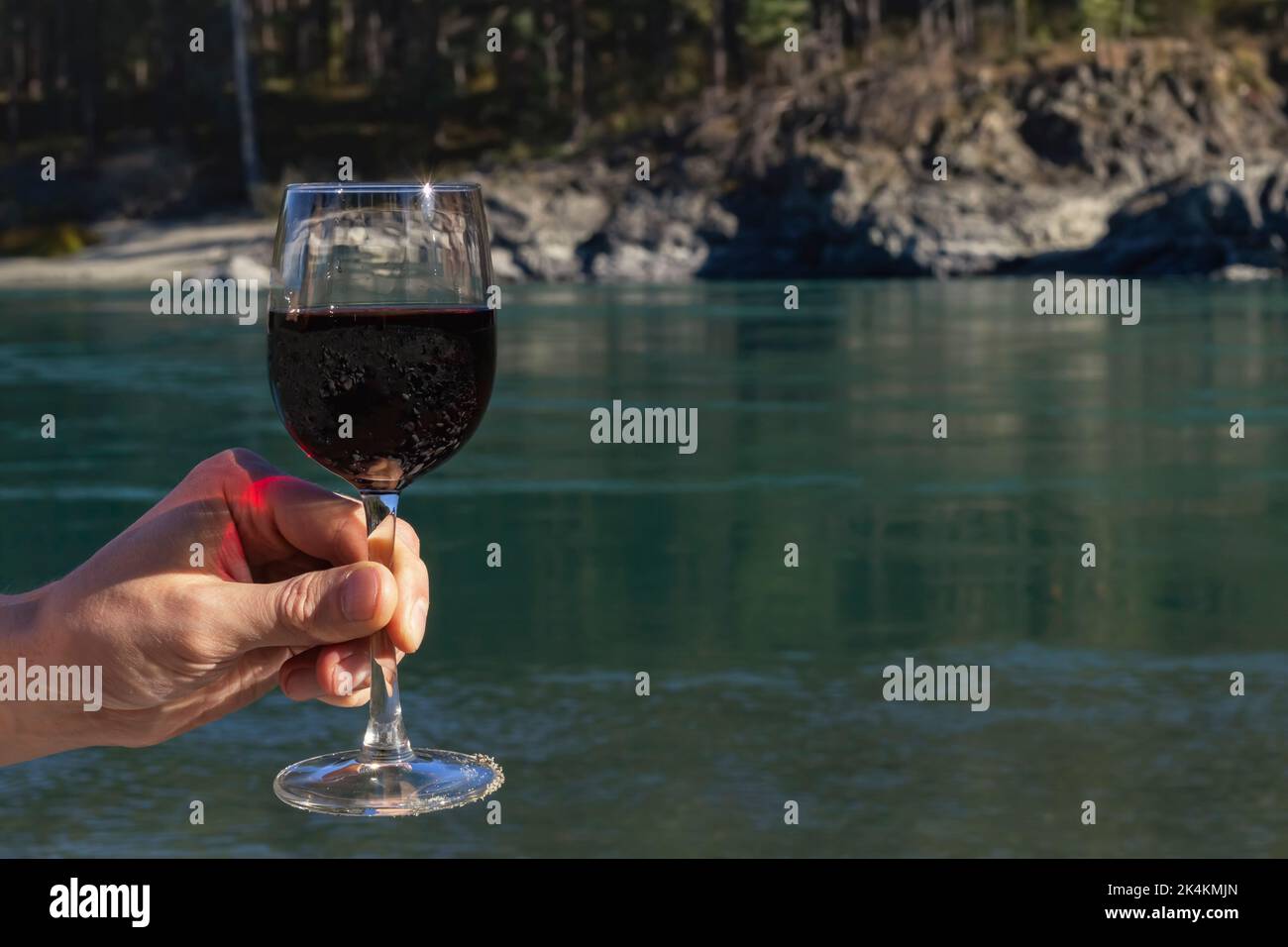 Frau Mann Hand mit einem Glas Rotwein auf dem Hintergrund des blauen Bergflusses am Strand.Konzepte Ernte und Weinherstellung, Urlaub, Reisen und Romantik in Stockfoto