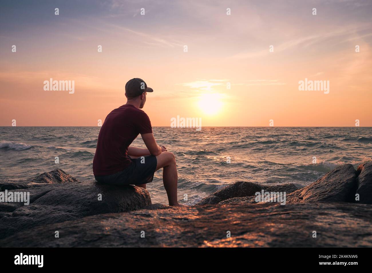 Rückansicht eines Mannes, der auf einem Felsen sitzt und den idyllischen Sonnenuntergang über dem Meer beobachtet. Stockfoto