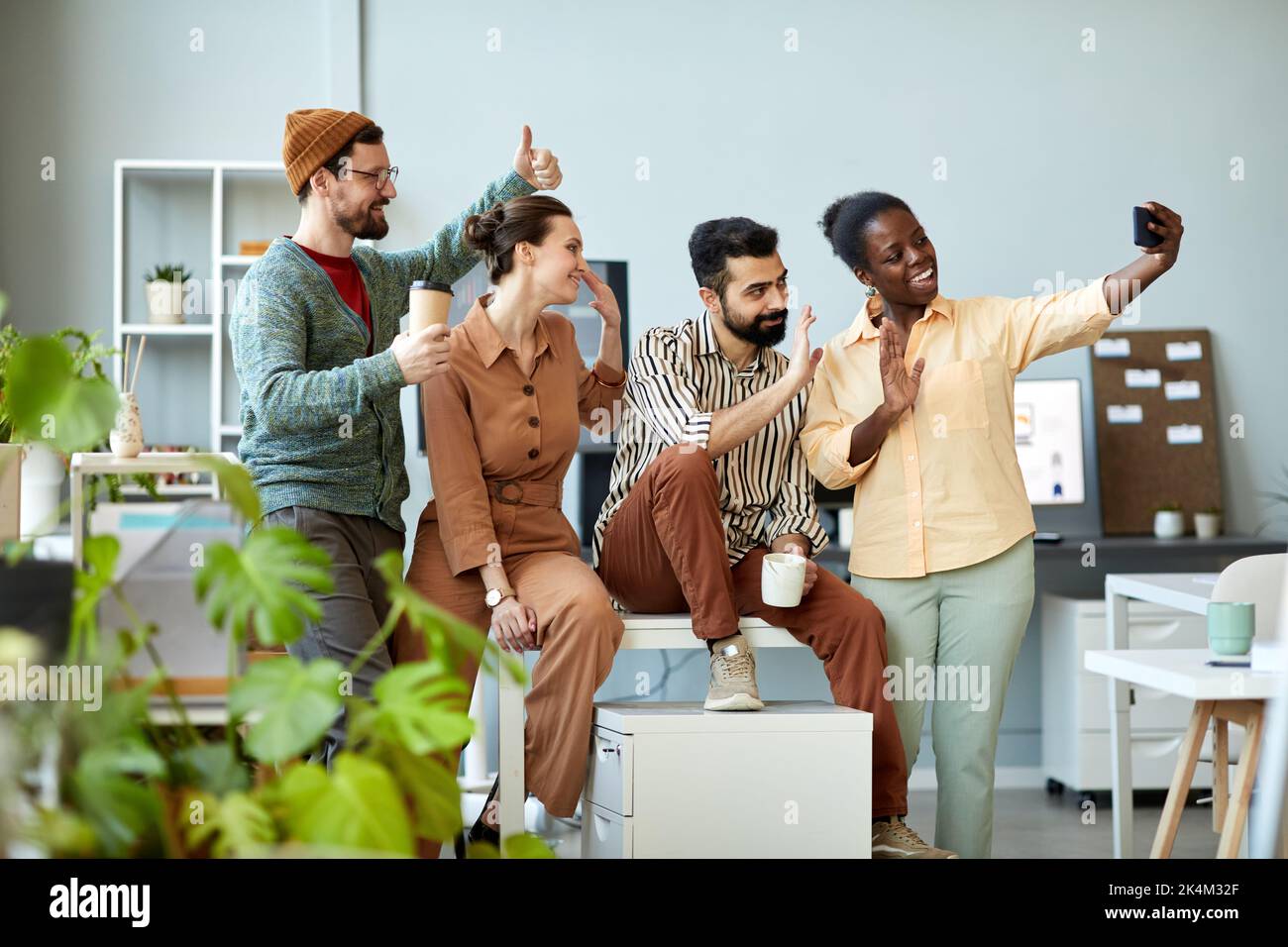 Seitenansicht von vier jungen, fröhlichen Mitarbeitern, die mit ihren Händen schwenken und lächelnd auf die Smartphone-Kamera blicken, während sie im Video-Chat kommunizieren Stockfoto