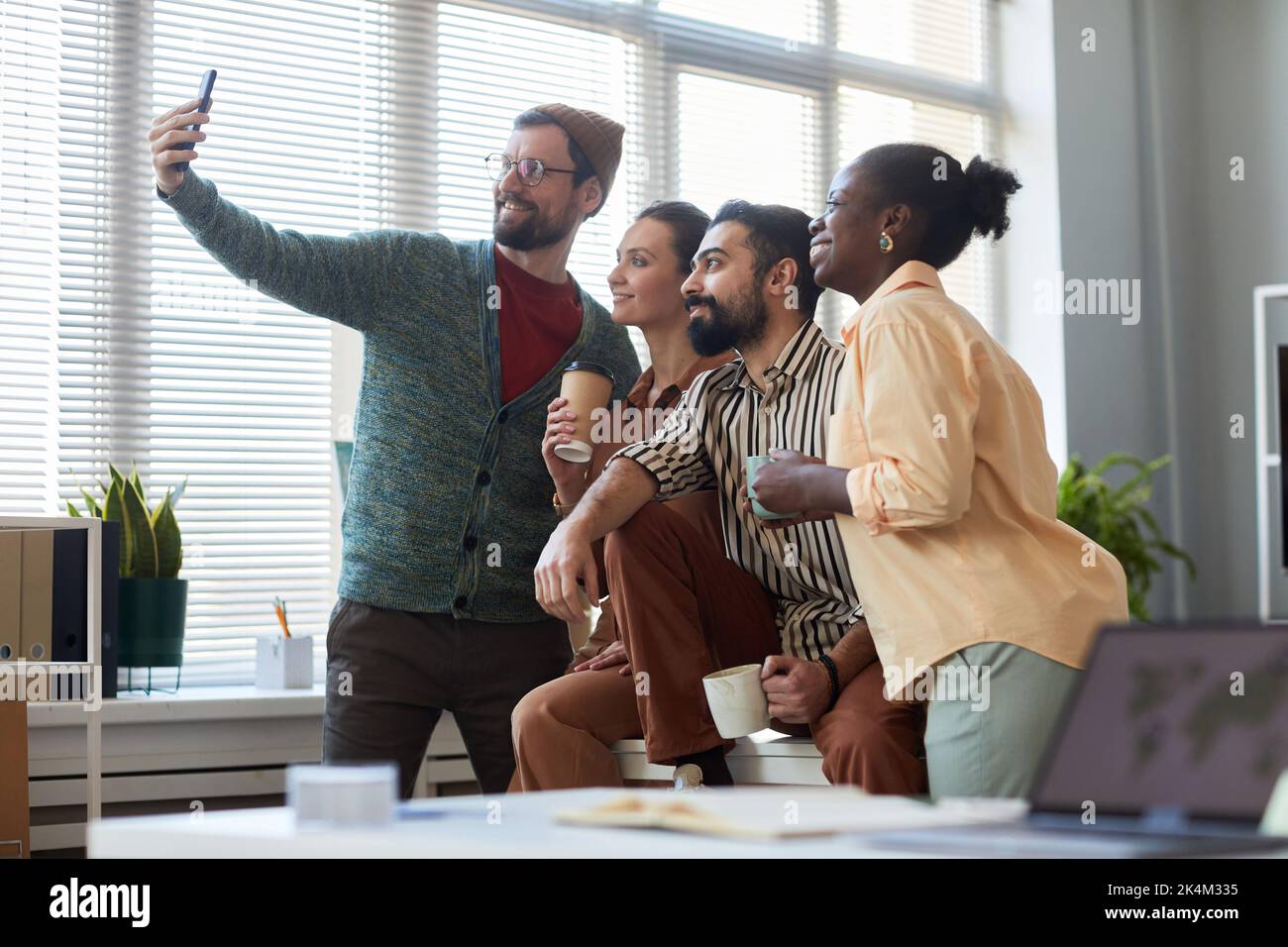 Junge, fröhliche Kollegen in smarter Casualwear blicken auf die Smartphone-Kamera, während sie am Arbeitsplatz im Openspace-Büro Selfie machen Stockfoto