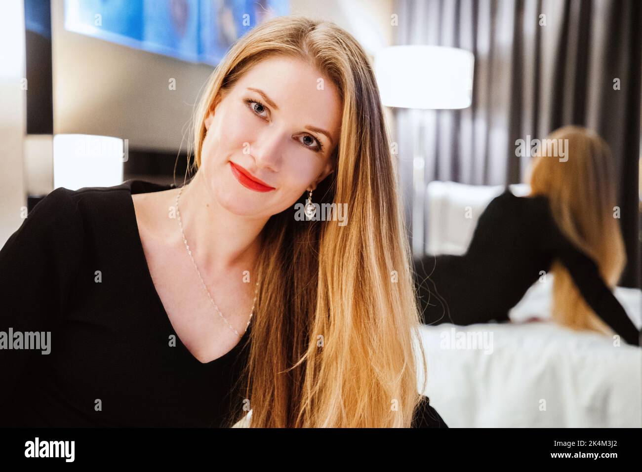 Hübsche Hündin mit langen blonden Haaren, in einem schwarzen Kleid, sitzend auf dem Bett, schaut uns an. Ihre Reflexion ist im Spiegel hinter ihr sichtbar Stockfoto