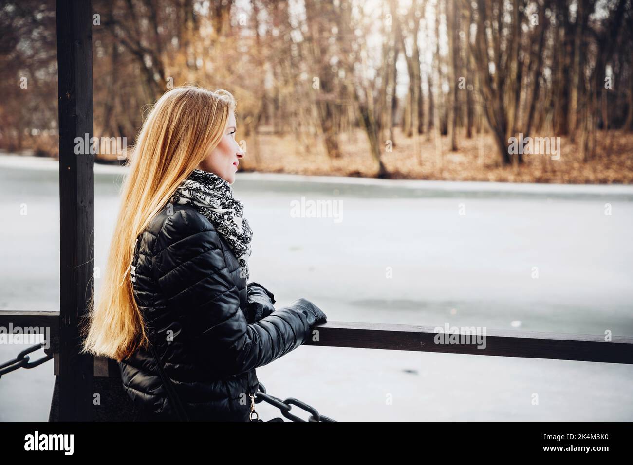 Das hübsche blonde Mädchen, das einen schwarzen Mantel, Handschuhe und Schal trägt, blickt über einen gefrorenen See; Bäume im Hintergrund Stockfoto