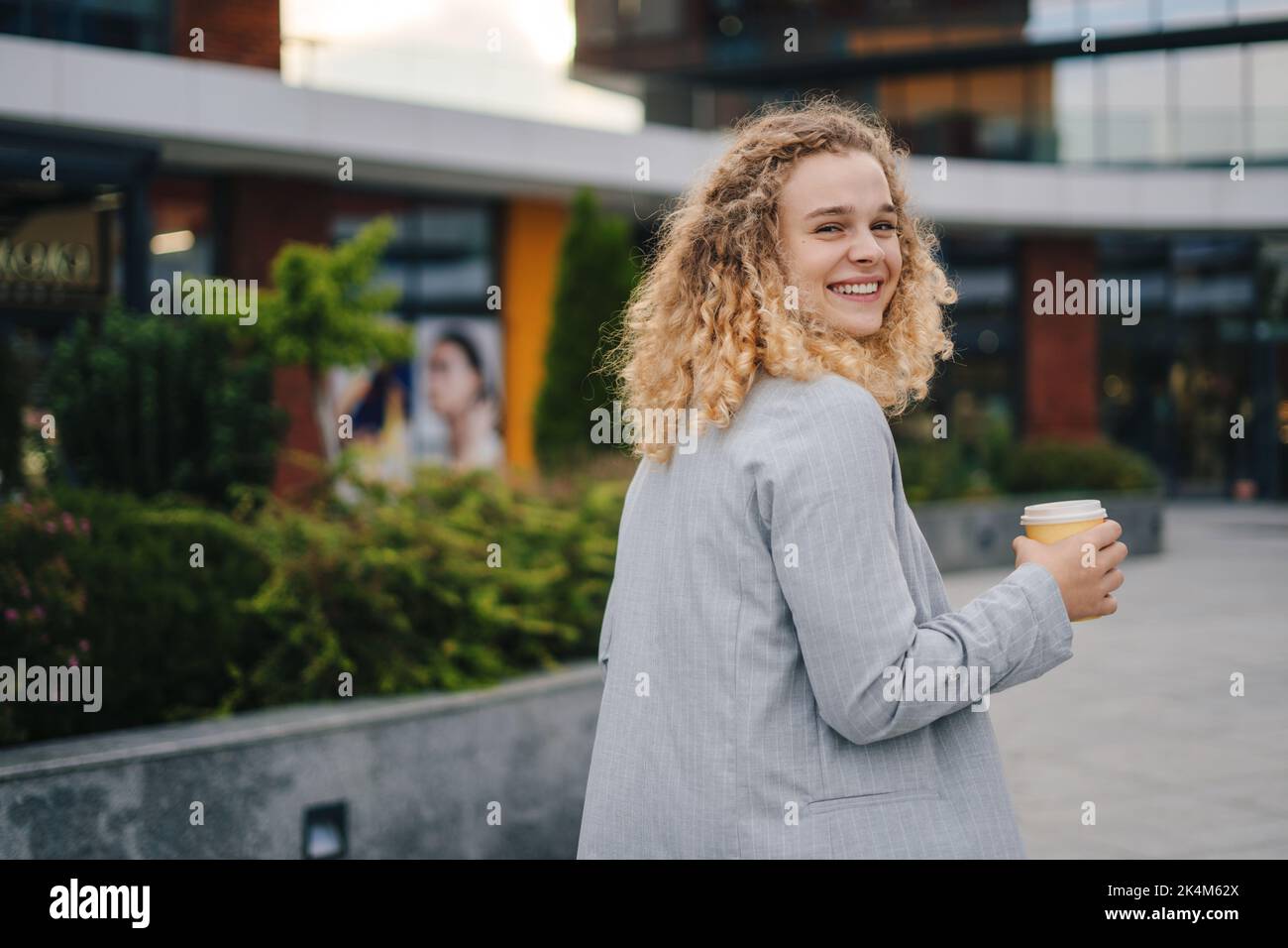 Foto einer attraktiven Studentin, die ihren Kopf zur Kamera dreht, während sie zum Eingang der Universität geht und Kaffee hält, um zu gehen und eine Stockfoto