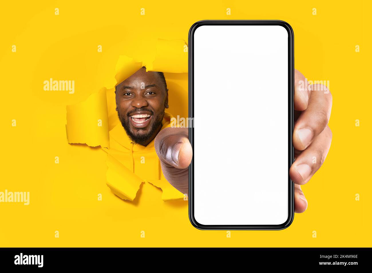 Empfehlung. Aufgeregt schwarzer Mann hält großes Smartphone mit leerem Bildschirm durch zerrissene gelbe Papierloch Stockfoto