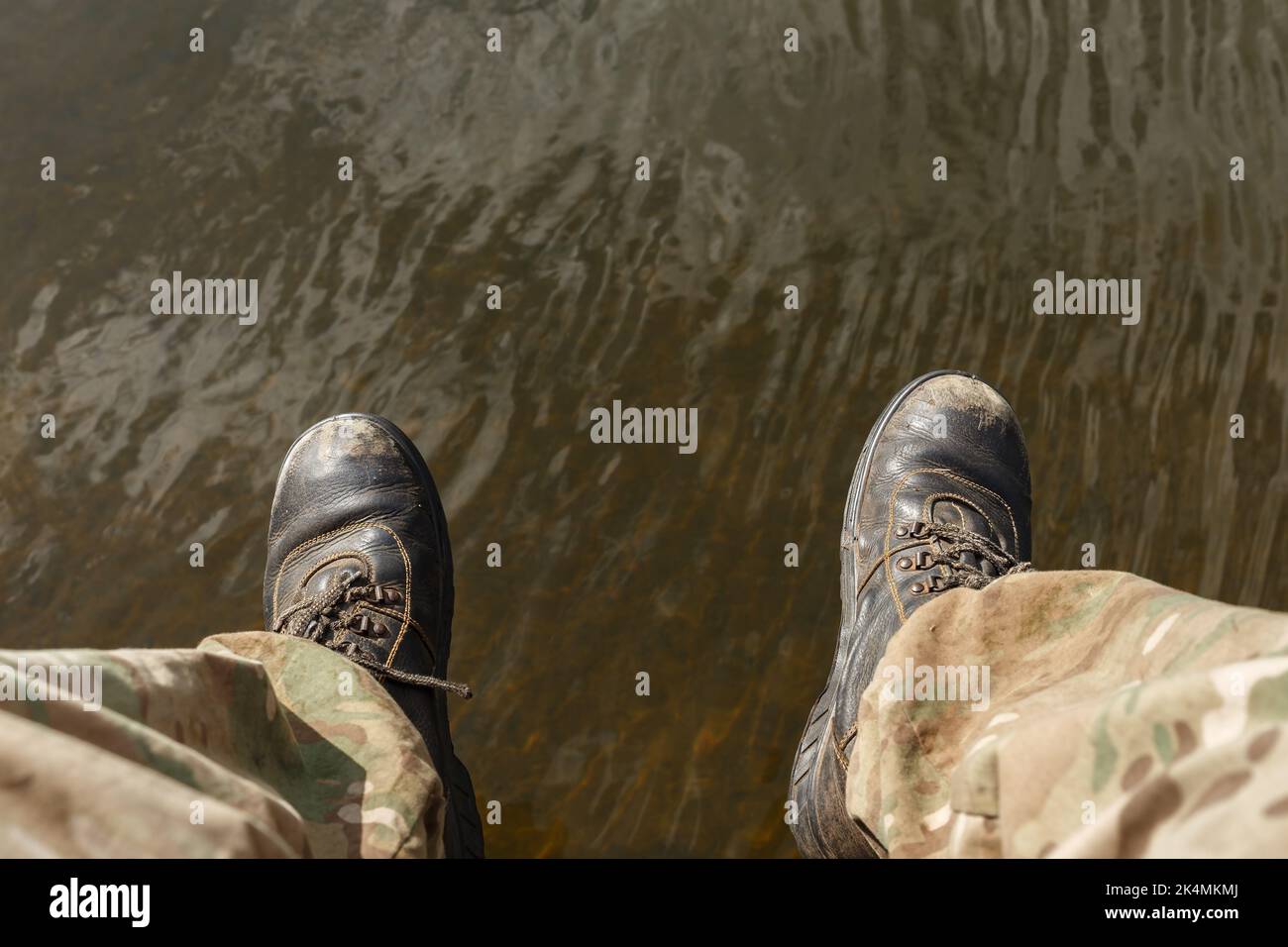 Niedriger Personenabschnitt über Wasser. Herrenbeine in Camouflage-Hosen und alten Stiefeln baumeln über dem Wasser. Stockfoto