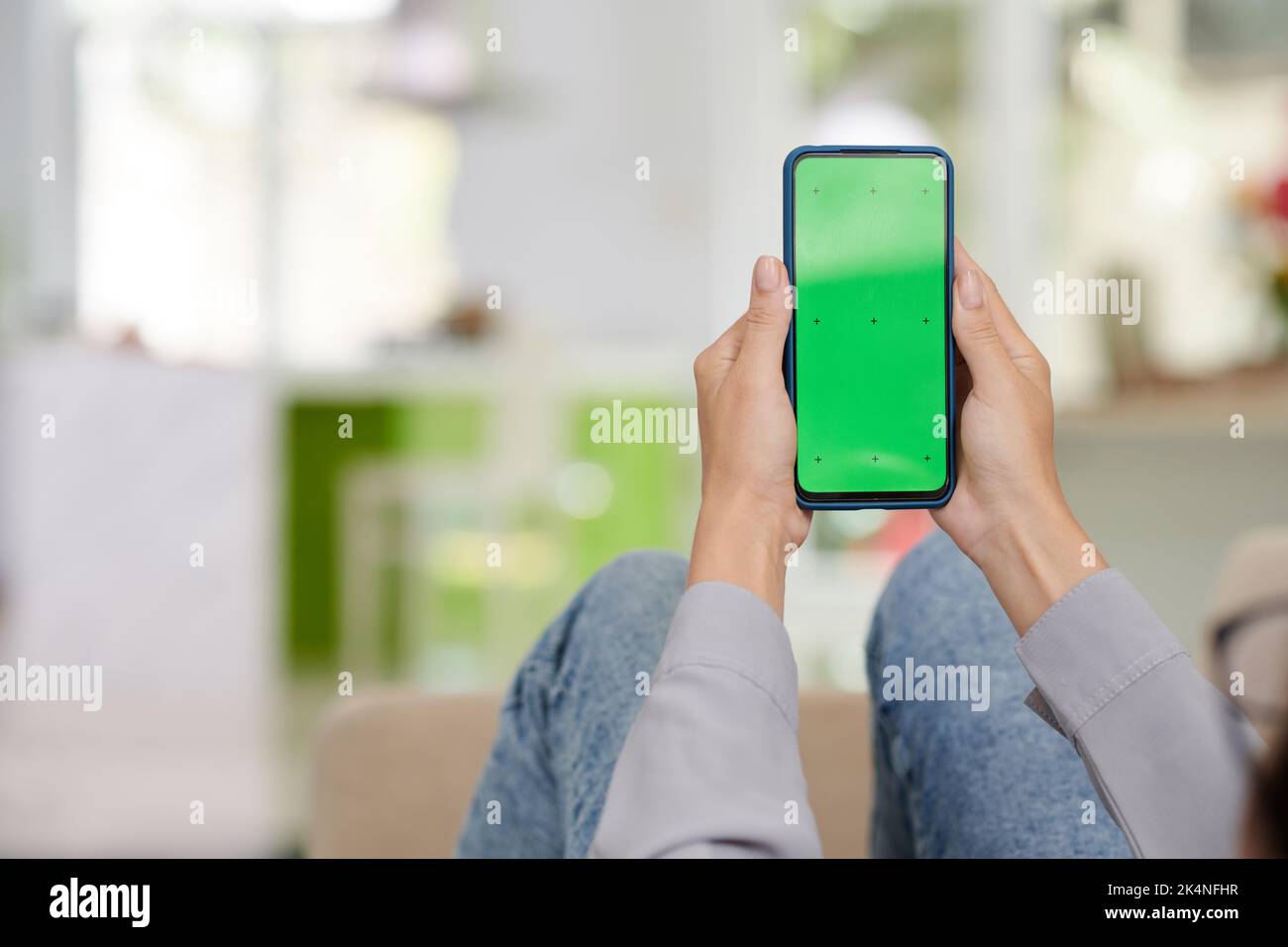 Smartphone mit leerem grünen Bildschirm, der von einer jungen Frau in einer legeren Kleidung gehalten wird, die vor der Kamera sitzt und eine neue mobile Anwendung verwendet Stockfoto