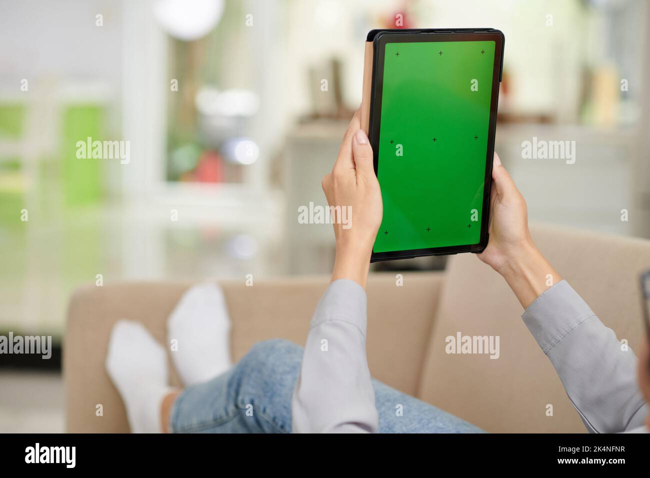 Nahaufnahme einer jungen modernen Frau in Casualwear, die ein Tablet mit leerem grünen Bildschirm hält, während sie sich auf der Couch vor der Kamera entspannt Stockfoto