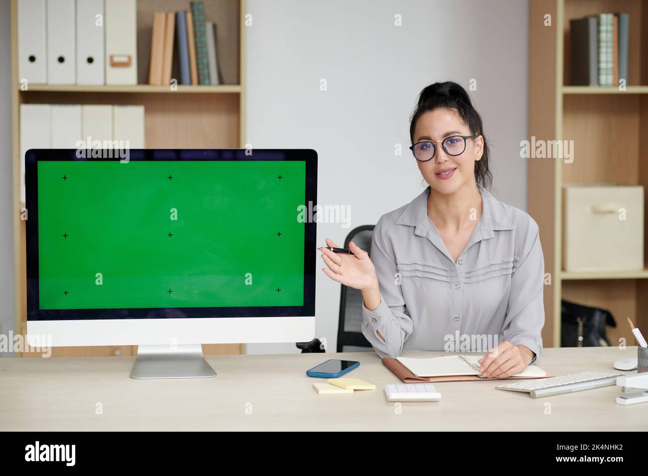 Junge Brünette Geschäftsfrau in Brillen und Casualwear macht Präsentation der neuen Anwendung, während sie auf leeren grünen Bildschirm zeigt Stockfoto