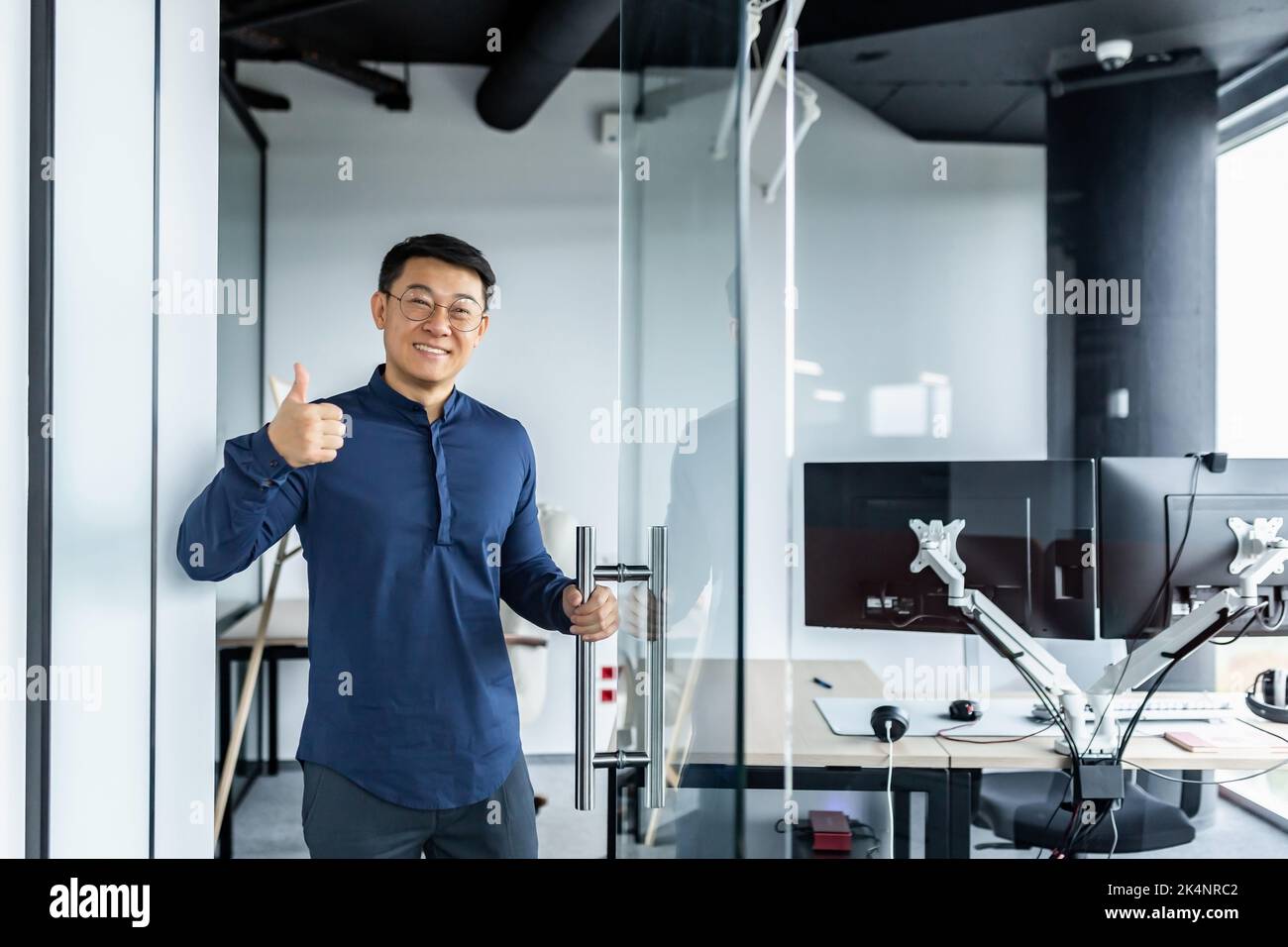 Porträt eines asiatischen Designers, eines lächelnden Mannes und eines bejahend haltenden Mannes, eines Geschäftsmanns, der mit mehreren Computern im verdrehten Bürogebäude arbeitete. Stockfoto