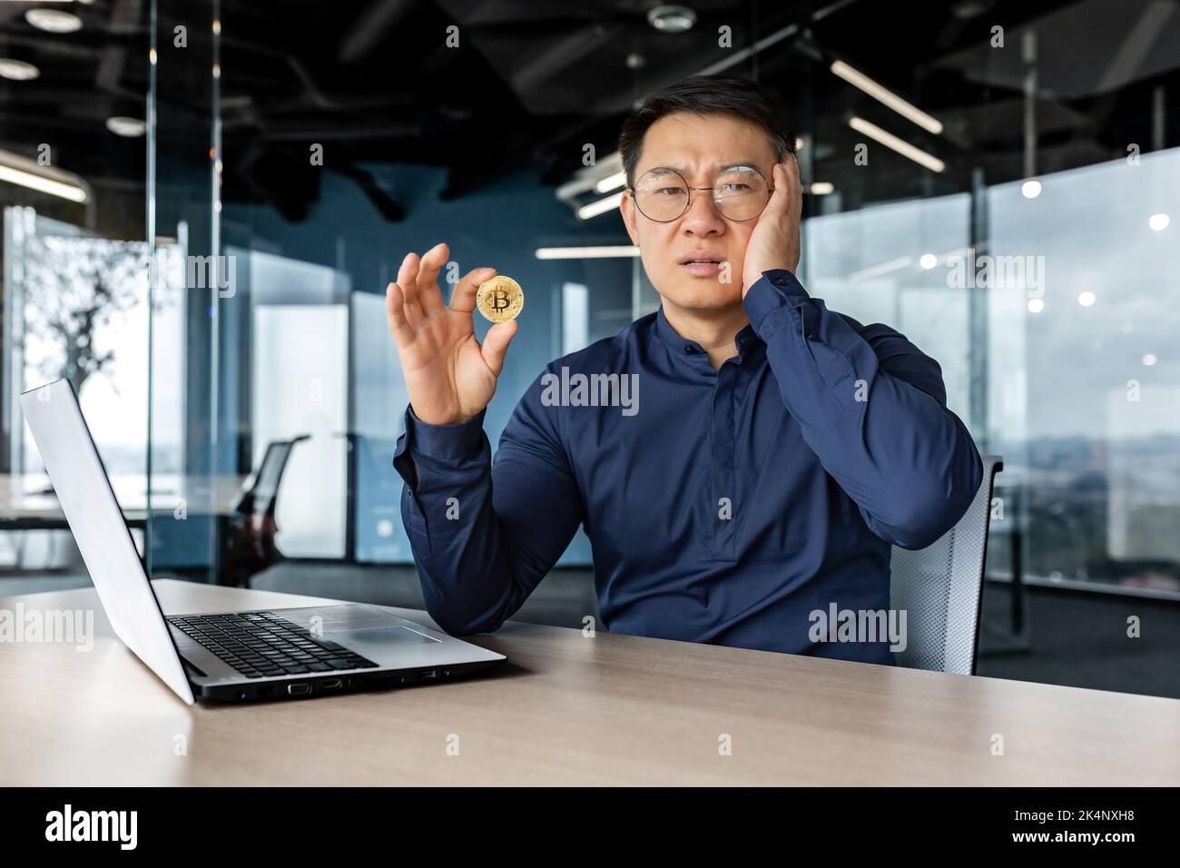 Unzufrieden und verwirrt in der Depression asiatischer Finanzinvestor, hält Münzgeld Krypto-Währung Bitcoin in den Händen, männlicher Geschäftsmann, der unzufrieden mit dem Wechselkurs arbeitet, Broker in Verzweiflung. Stockfoto