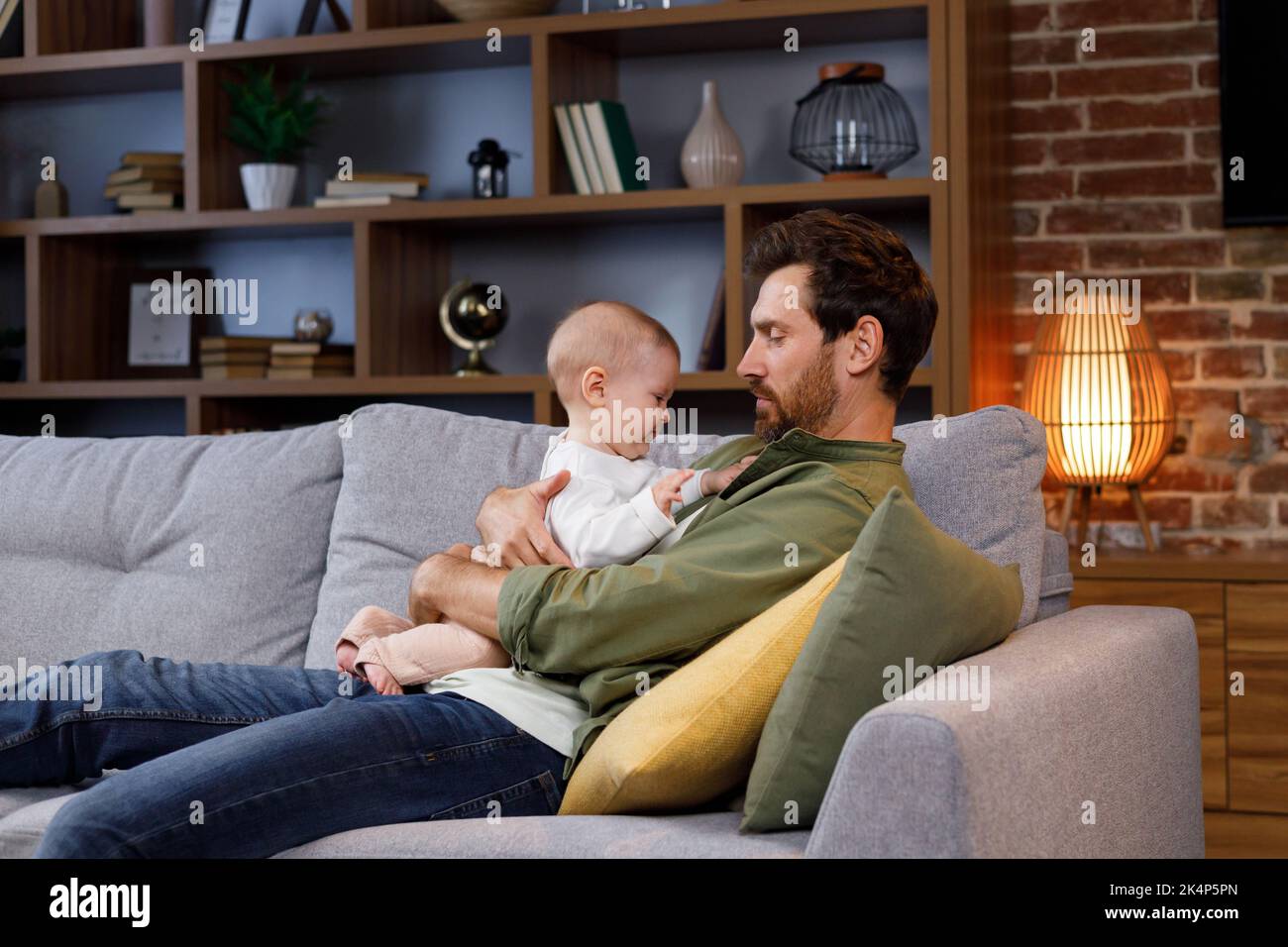 Papa spielt mit seinem kleinen Baby auf der Couch in einer gemütlichen Wohnung. Väterliche Liebe. Vater umarmt und küsst seine kleine Tochter. Stockfoto