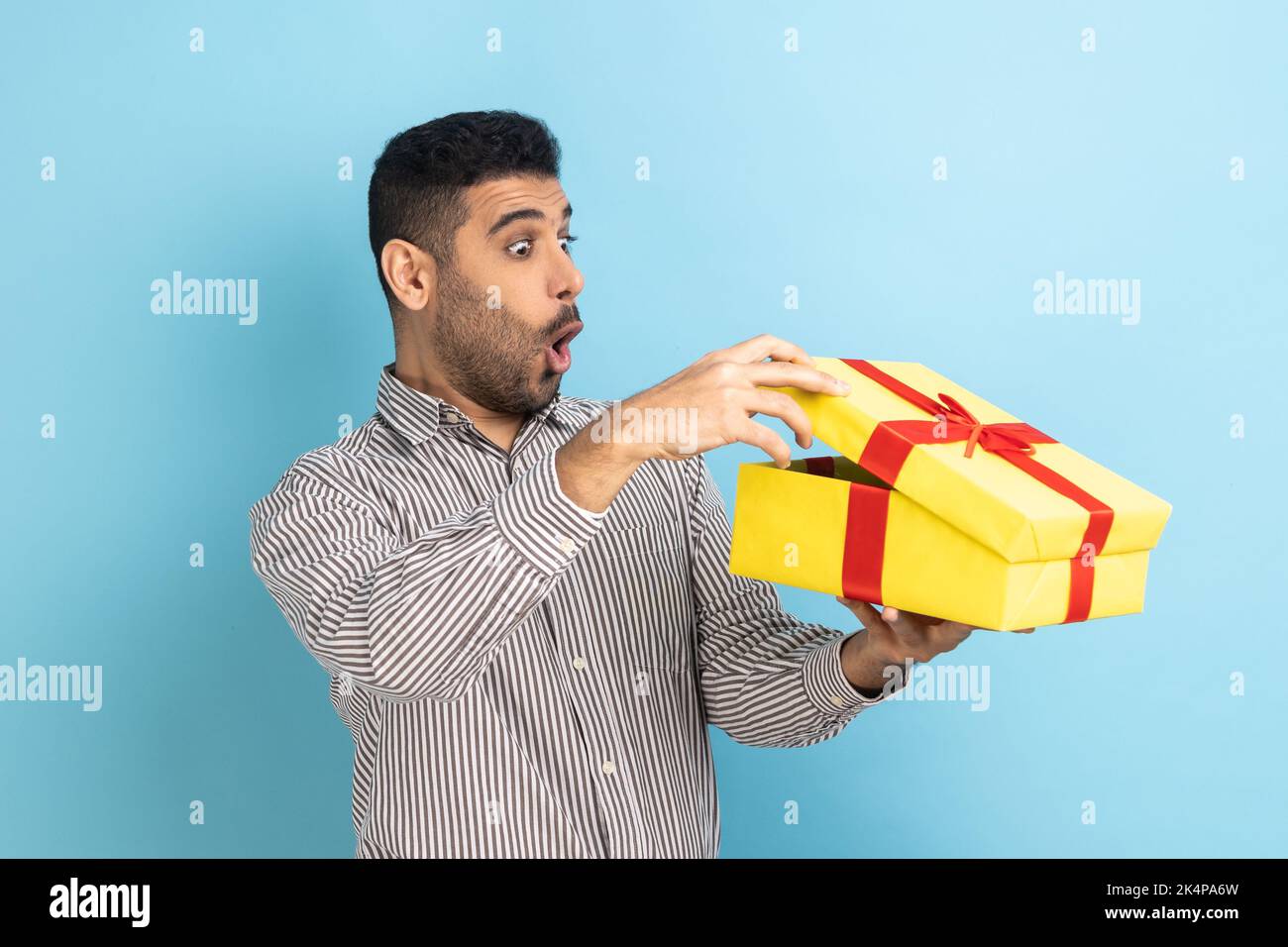 Porträt eines überraschten, neugierigen Geschäftsmannes, der in einer Geschenkschachtel schaut, das Geschenk öffnet und mit Interesse in das Innere blickt, in einem gestreiften Hemd. Innenaufnahme des Studios isoliert auf blauem Hintergrund. Stockfoto