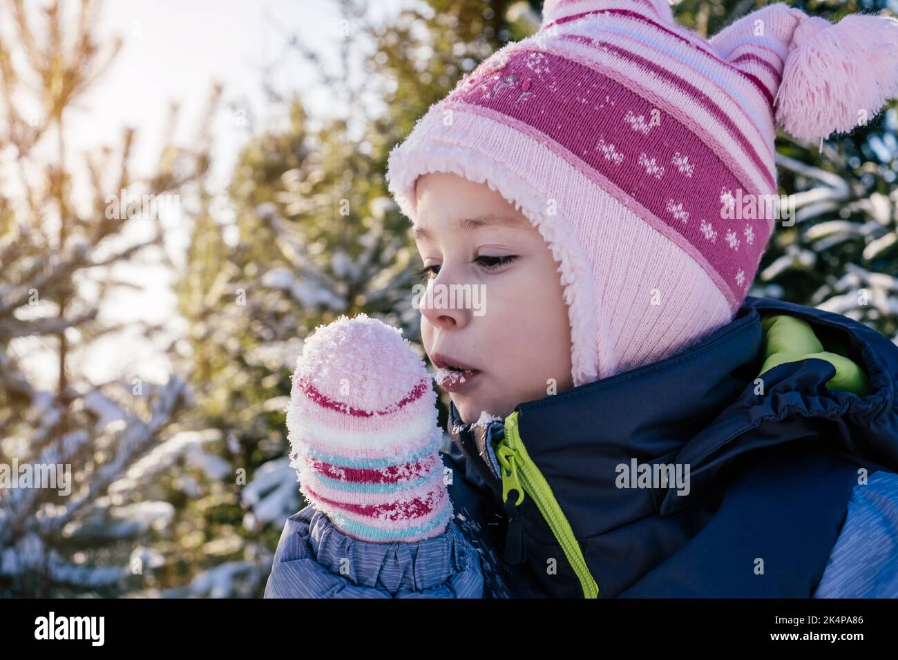 Kleines Mädchen 3-4 Jahre alt in Winterkleidung Overalls, Hut und Fäustlinge, steht im Freien gegen schneebedeckte Pinien und Tannen und frisst Schnee an sonnigen Stockfoto