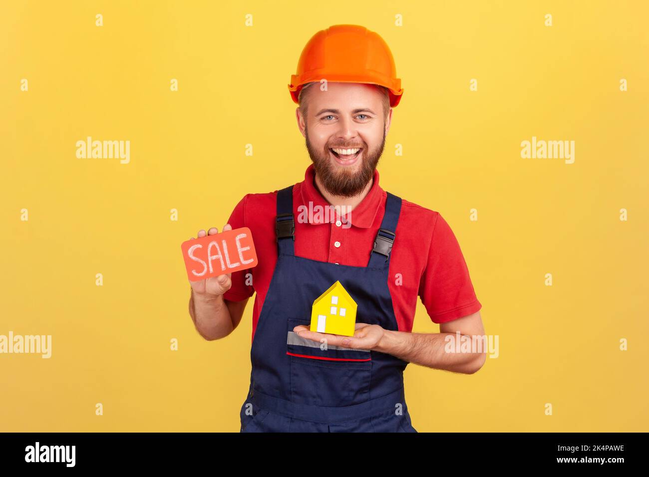 Porträt eines begeisterten Bauherrn in blauer Uniform und Schutzhelm mit Verkaufskarte und Papierhaus, große Slae für Immobilien. Innenaufnahme des Studios isoliert auf gelbem Hintergrund. Stockfoto