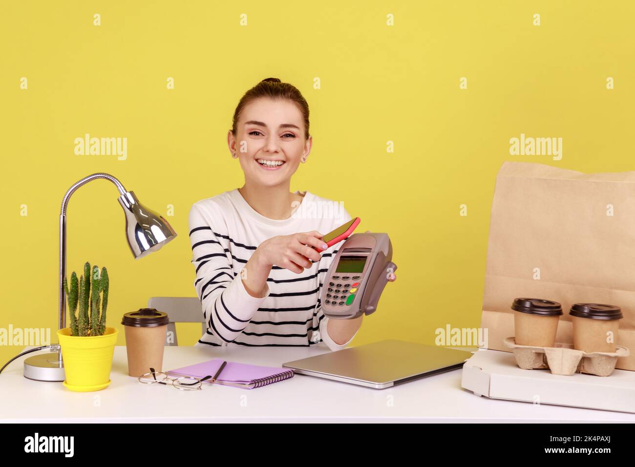 Glückliche junge Frau, die am Arbeitsplatz mit einem mobilen Smartphone sitzt und per Pay-Pass Pos. kontaktlos, nfc-Technologie auf dem Handy und Zahlung zahlt. Studio-Innenaufnahme isoliert auf gelbem Hintergrund Stockfoto