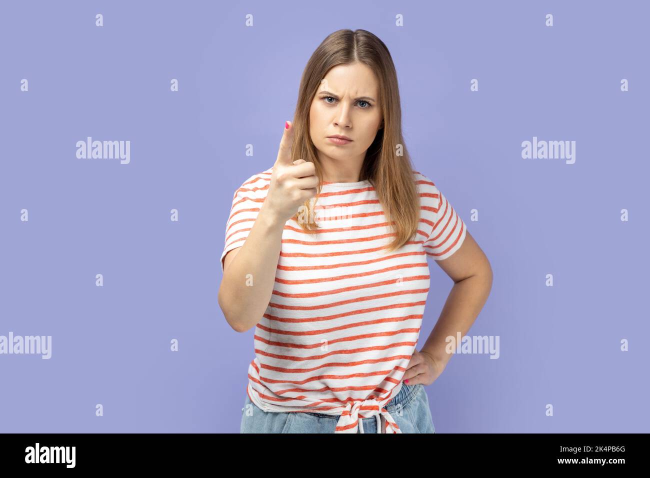 Porträt einer blonden Frau mit gestreiftem T-Shirt, der mit dem Finger auf die Kamera zeigt und mit unzufriedenem, verdächtigem Ausdruck vor Problemen warnt. Innenaufnahme des Studios isoliert auf violettem Hintergrund. Stockfoto