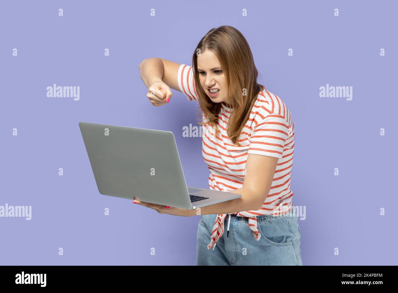 Porträt einer wütenden, aggressiven, blonden Frau mit gestreiftem T-Shirt, die am Laptop arbeitet, Online-Gespräche führt, auf dem Notebook-Display boxt. Innenaufnahme des Studios isoliert auf violettem Hintergrund. Stockfoto