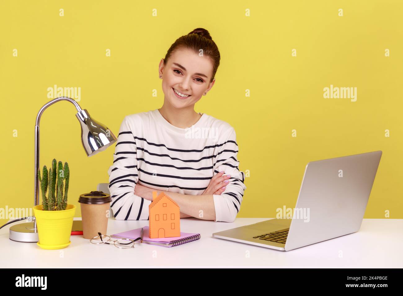 Porträt einer glücklichen Frau am Arbeitsplatz mit einem kleinen Papierhaus, das mit einem Lächeln auf die Kamera blickt und von ihrer eigenen Wohnung träumt. Studio-Innenaufnahme isoliert auf gelbem Hintergrund. Stockfoto